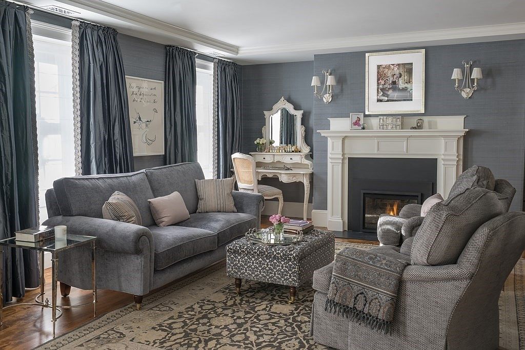 This is an interior shot of a living room featuring a gray sofa with decorative pillows, a patterned ottoman, and a gray armchair. A fireplace with a white mantel is visible in the background, along with elegant wall sconces and framed artwork. The room is decorated in a sophisticated, neutral color palette with gray walls and curtains, creating a cozy and inviting atmosphere.