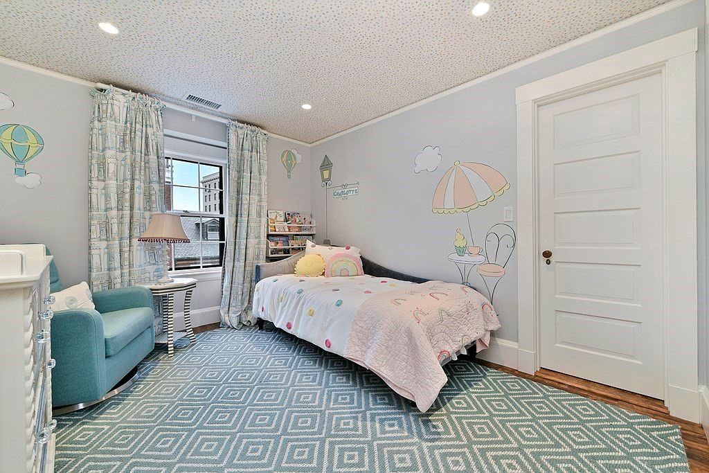 This is a charming children's bedroom featuring light blue walls adorned with whimsical drawings and a patterned ceiling. The room includes a twin bed with a colorful quilt, a cozy teal armchair, and a patterned blue and white rug. Natural light streams in through a window with patterned curtains, creating a bright and inviting space.