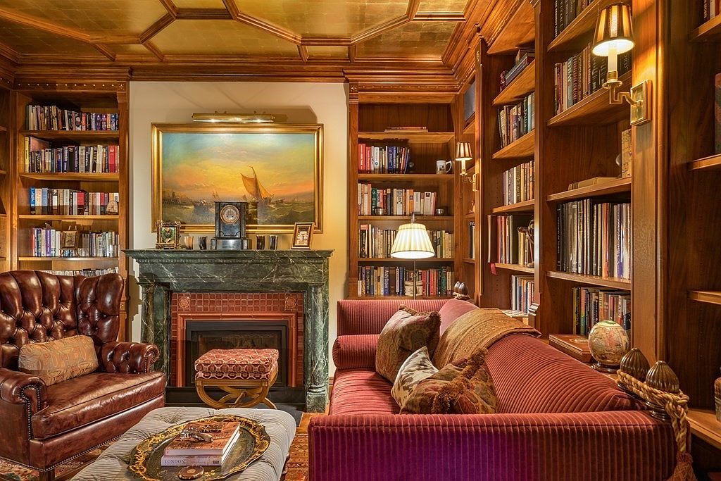 This is a cozy and elegant living room featuring a fireplace with a green marble surround, flanked by built-in bookshelves filled with books. A comfortable leather armchair and a plush red sofa provide ample seating, while a large painting hangs above the fireplace, adding a touch of sophistication. The room exudes a warm and inviting atmosphere, perfect for relaxation and reading.
