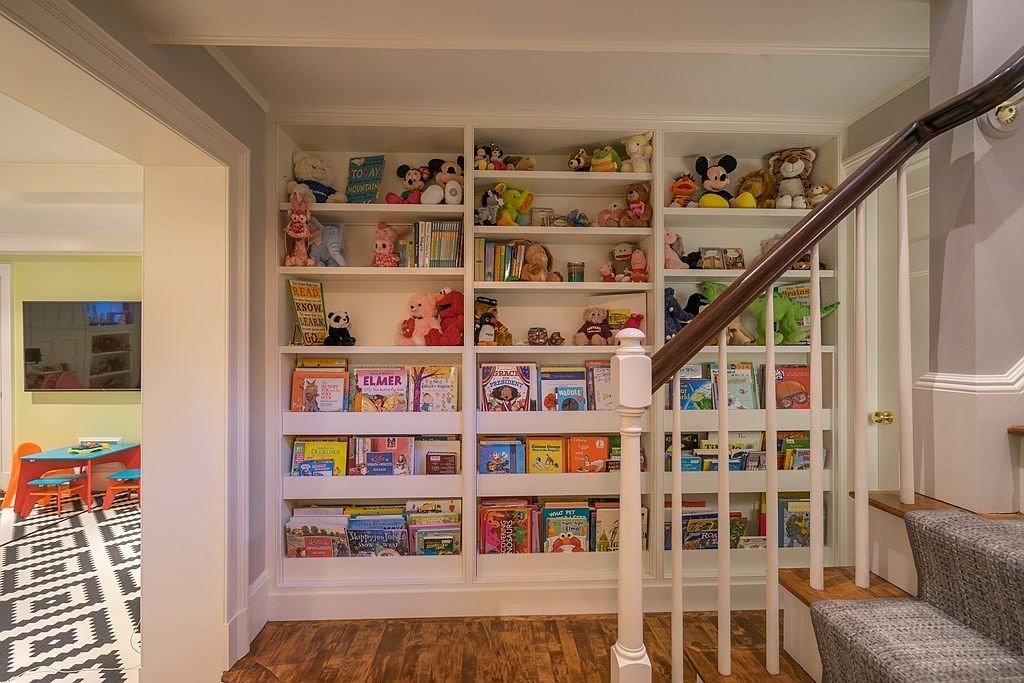 This interior shot showcases a hallway with a built-in bookshelf filled with books and stuffed animals, creating a cozy and inviting atmosphere. A staircase with white railings and a wooden handrail is visible to the right, leading to another level of the home. The flooring is hardwood, and the adjacent room features a playful black and white patterned floor, adding a touch of whimsy.