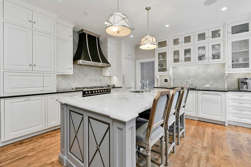 This is a bright and spacious kitchen featuring white cabinetry, a large center island with a marble countertop, and stainless steel appliances. The kitchen is well-lit with pendant lighting and recessed lights, and the hardwood floors add warmth to the space. The overall impression is one of luxury and functionality, perfect for a modern home.