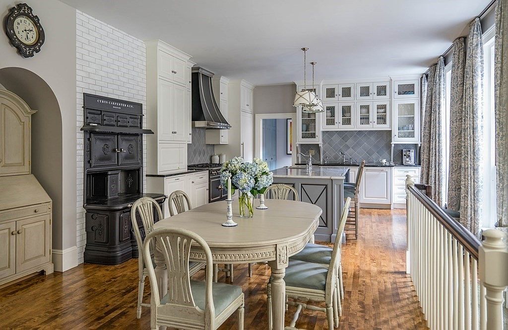 This is an interior shot of a well-appointed kitchen and dining area. The kitchen features white cabinetry, a gray island, and stainless steel appliances, complemented by a gray backsplash. The dining area includes a light-colored wooden table with matching chairs, creating a cohesive and elegant space.