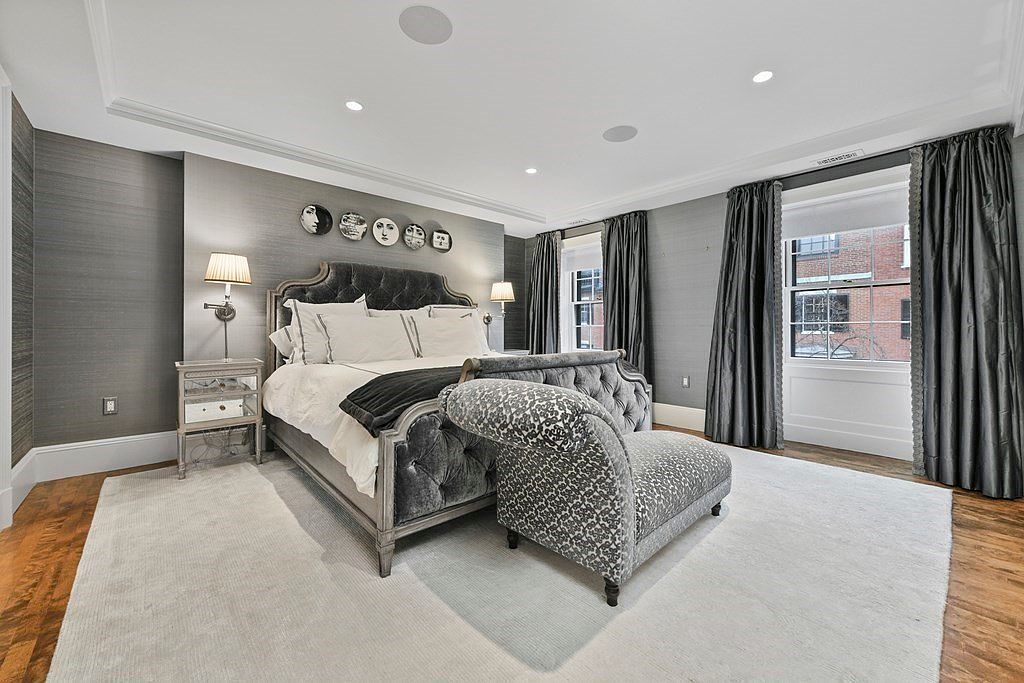 This is a primary bedroom featuring a luxurious upholstered bed with white linens and a dark throw, complemented by a patterned chaise lounge. The room is decorated in a sophisticated gray palette with textured wallpaper, accented by modern wall art and elegant window treatments. A plush white rug covers hardwood floors, adding warmth and comfort to the space.