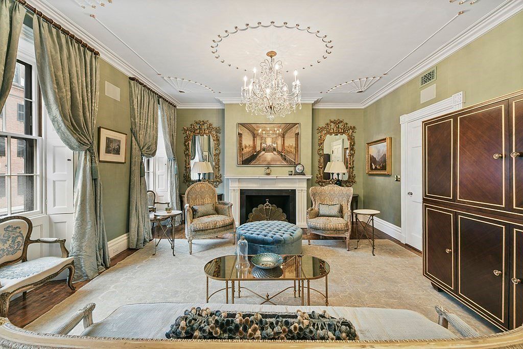 This is an elegant living room featuring a fireplace with a decorative mantel, flanked by ornate mirrors and comfortable armchairs. A crystal chandelier hangs from the detailed ceiling, and the room is furnished with a plush ottoman, a glass-topped coffee table, and luxurious drapes. The overall impression is one of refined sophistication and classic design.