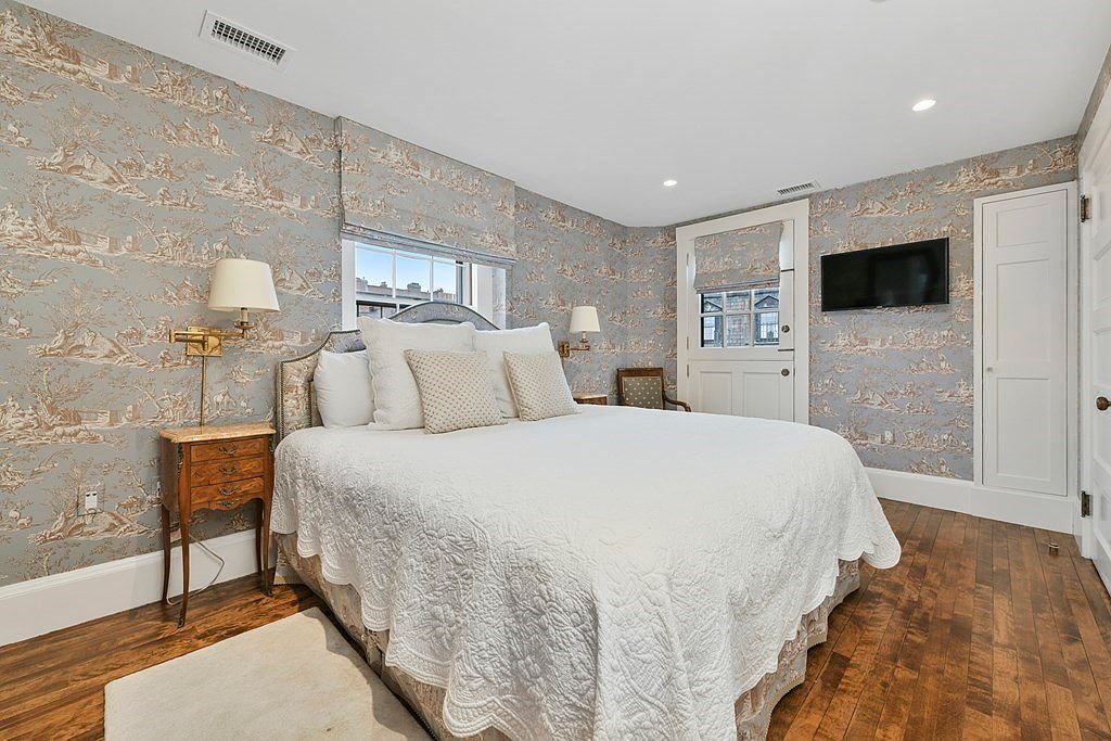 This is a primary bedroom featuring a queen-sized bed with white bedding, a wooden nightstand with a lamp, and walls adorned with patterned wallpaper. The room has hardwood floors and a flat-screen TV mounted on the wall. The overall style is traditional and cozy.
