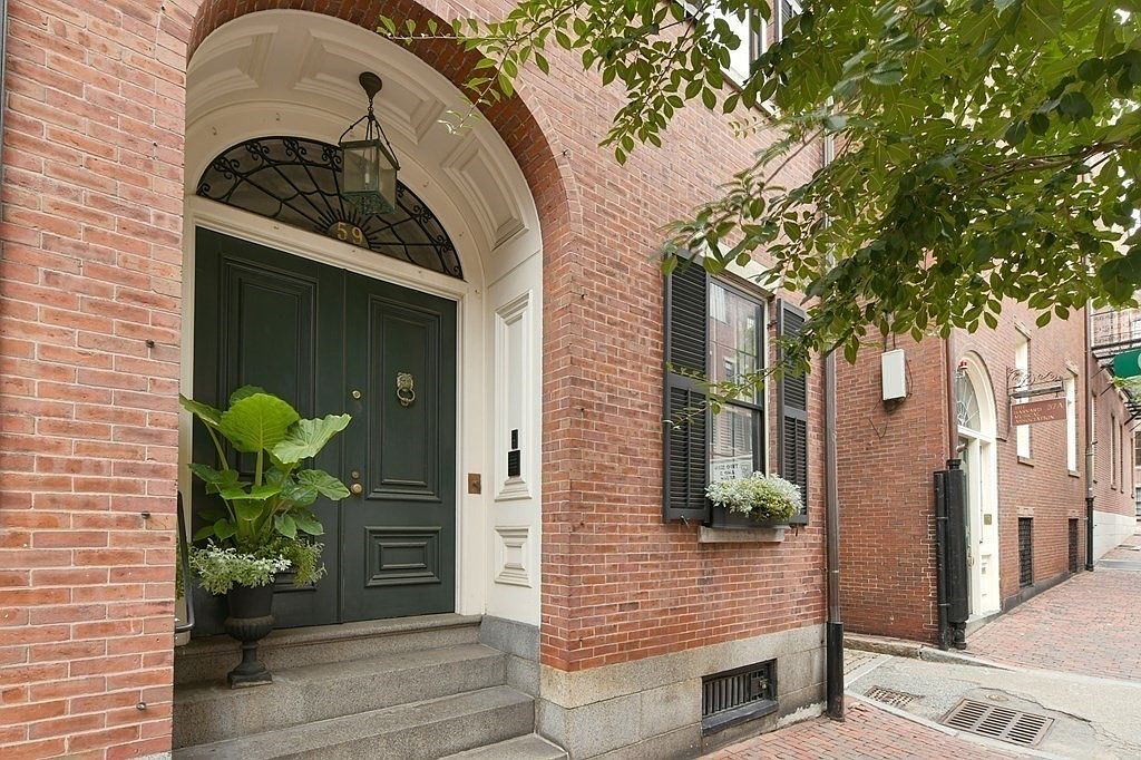 This image showcases the elegant entryway of a brick townhouse. A dark green double door is framed by a white archway and topped with a decorative wrought iron detail and hanging lantern. A potted plant adds a touch of greenery, while the brick facade and window with shutters contribute to the building's classic charm.