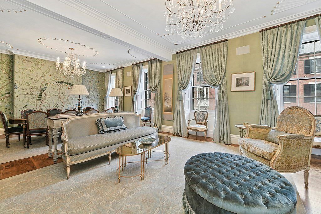 This elegant living room features a sophisticated blend of classic and refined elements. The room is furnished with a plush sofa, ornate armchairs, and a tufted ottoman, all set upon an intricately patterned rug. The walls are adorned with decorative wallpaper and complemented by flowing curtains, creating a luxurious and inviting atmosphere.