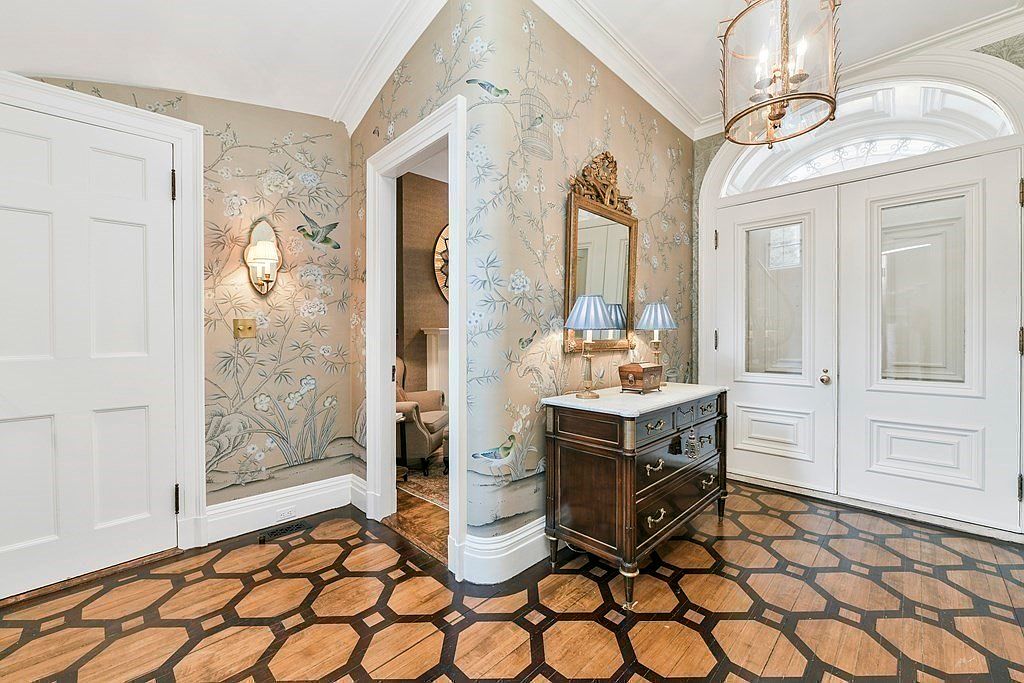 This is an interior shot of a grand hallway featuring patterned wood flooring and elegant wallpaper with bird and floral motifs. A dark wood console table with a marble top is adorned with lamps and a mirror, adding to the sophisticated ambiance. The white doors and trim provide a crisp contrast to the warm tones of the floor and wallpaper, creating a welcoming and luxurious entryway.