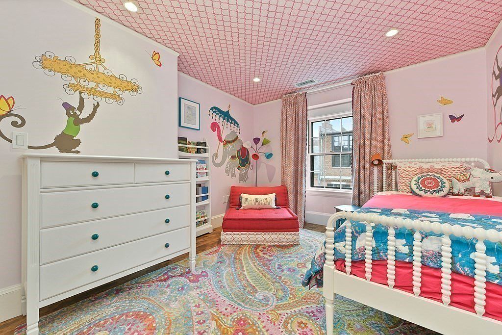 This is a charming children's bedroom featuring whimsical decor. A white dresser with teal knobs sits against a wall adorned with a monkey mural, while a colorful paisley rug covers the floor. The room also includes a white spindle bed with pink and blue bedding, and a red lounge chair, creating a playful and inviting atmosphere.