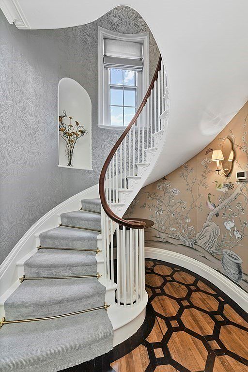 This image showcases a beautifully designed spiral staircase with carpeted steps and a wooden handrail. The walls are adorned with elegant wallpaper, and a window provides natural light. The flooring at the base of the stairs features a unique geometric pattern, adding a touch of sophistication to the space.
