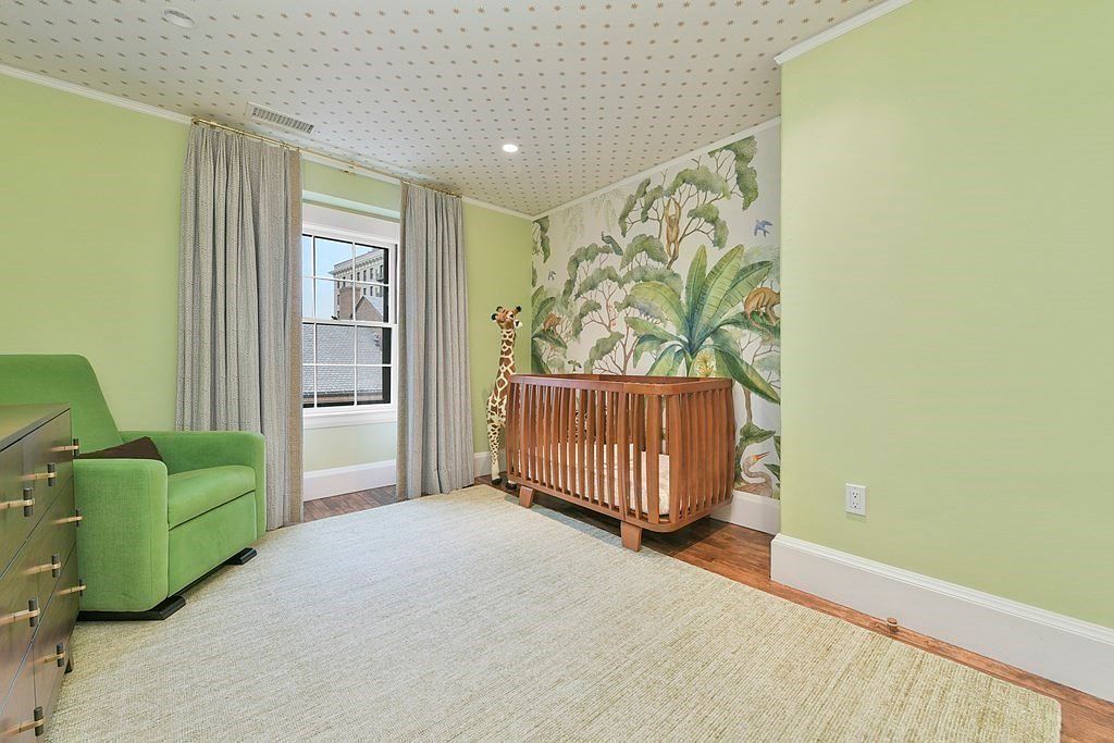 This is a charming nursery featuring light green walls, a patterned ceiling, and a jungle-themed accent wall. A wooden crib sits against the accent wall, complemented by a giraffe plush toy. The room also includes a comfortable green armchair and a dresser, creating a cozy and inviting space for a child.