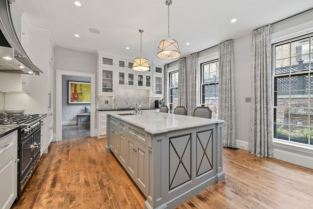 This is a well-lit kitchen featuring a large central island with a marble countertop and gray cabinetry with decorative X panels. The kitchen also includes white upper cabinets, some with glass fronts, stainless steel appliances, and hardwood flooring. Two pendant lights hang above the island, and large windows with patterned curtains provide natural light.