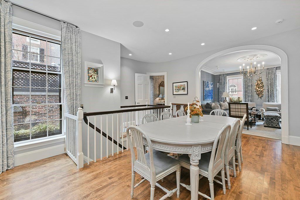 This is an interior shot of a dining room featuring a large, oval-shaped dining table with white painted finish and matching chairs with light gray cushions. The room has hardwood floors and is well-lit with recessed lighting and natural light from a large window. The dining room opens into a living room, creating an open-concept feel.
