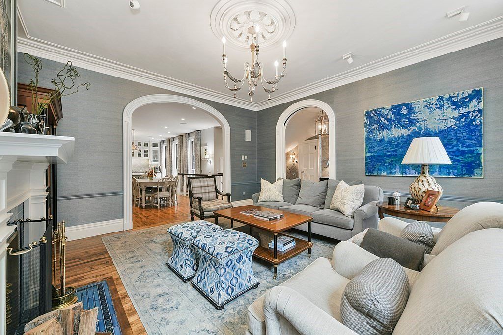 This is an interior shot of a living room featuring a gray sofa, a white sofa, and a wooden coffee table. The room has a sophisticated design with blue wallpaper, white trim, and a decorative chandelier. Arched doorways lead to other rooms, enhancing the open and inviting atmosphere.