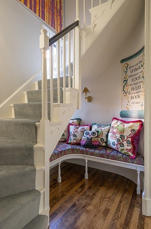 This image showcases a charming hallway with carpeted stairs and a cozy reading nook beneath the staircase. The nook features a curved bench adorned with colorful pillows, creating an inviting space. A wall decal with a Dr. Seuss quote adds a whimsical touch, enhancing the home's character.