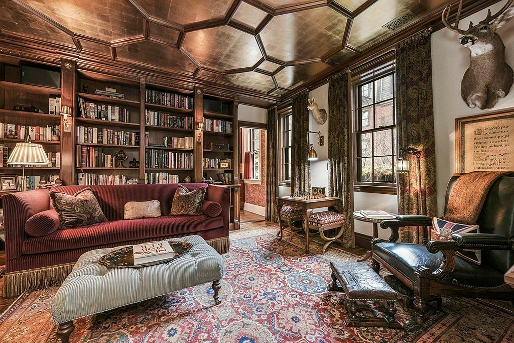 This is a luxurious and stately home office or study, featuring a large built-in bookcase filled with books, a comfortable red sofa, and a patterned rug. The room is well-lit by natural light from the windows, which are framed by elegant curtains, and the ceiling has a unique geometric design. The overall impression is one of sophistication and comfort, perfect for a discerning buyer.