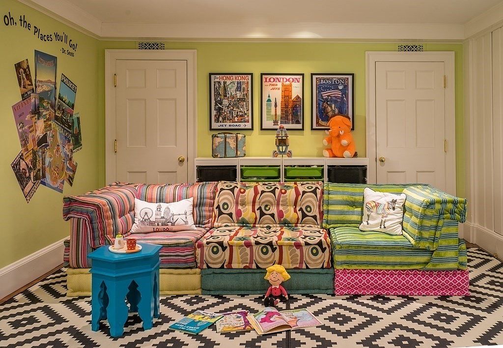 This vibrant living room features a playful and eclectic design, perfect for a family or children's space. The room is anchored by a modular sofa with colorful striped and patterned cushions, complemented by a whimsical blue side table and a black and white geometric rug. Travel-themed posters and a collection of books and toys add to the room's cheerful and inviting atmosphere.