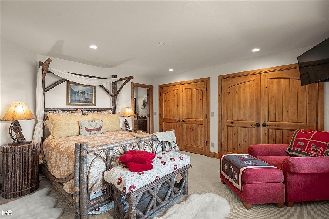 This is a cozy primary bedroom featuring a unique four-poster bed made of natural wood, adorned with soft bedding and decorative pillows. The room includes rustic-style furniture, such as a wooden bench and a side table, complemented by a plush rug. The overall aesthetic is warm and inviting, creating a comfortable retreat.