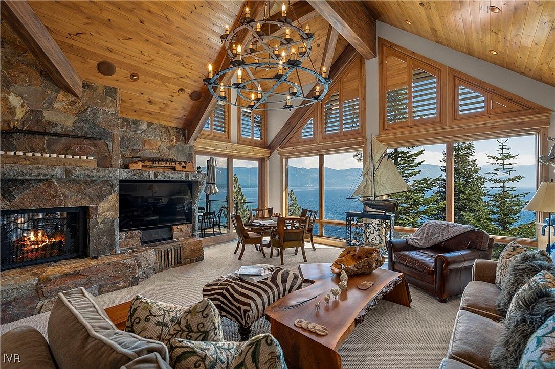 This is a luxurious living room featuring a stone fireplace with a mounted television, large windows offering a scenic lake view, and comfortable seating arrangements. The room is decorated with a rustic-chic style, incorporating natural wood elements and a unique chandelier, creating a warm and inviting atmosphere. The space is well-lit and appears spacious, perfect for relaxation and entertaining.