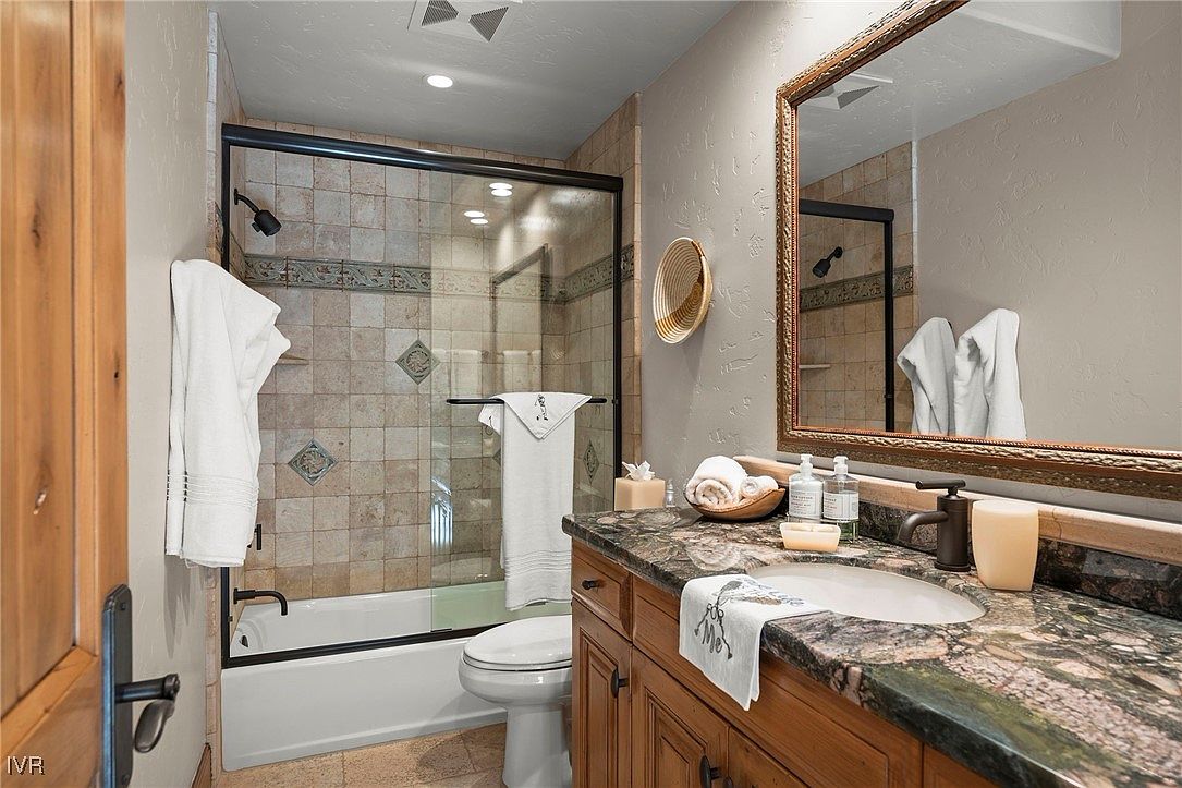 This is a bathroom featuring a shower/tub combination with a glass enclosure and tiled walls. The vanity has a granite countertop and wooden cabinets, with a decorative mirror above. The overall style is traditional and well-maintained, creating a comfortable and functional space.