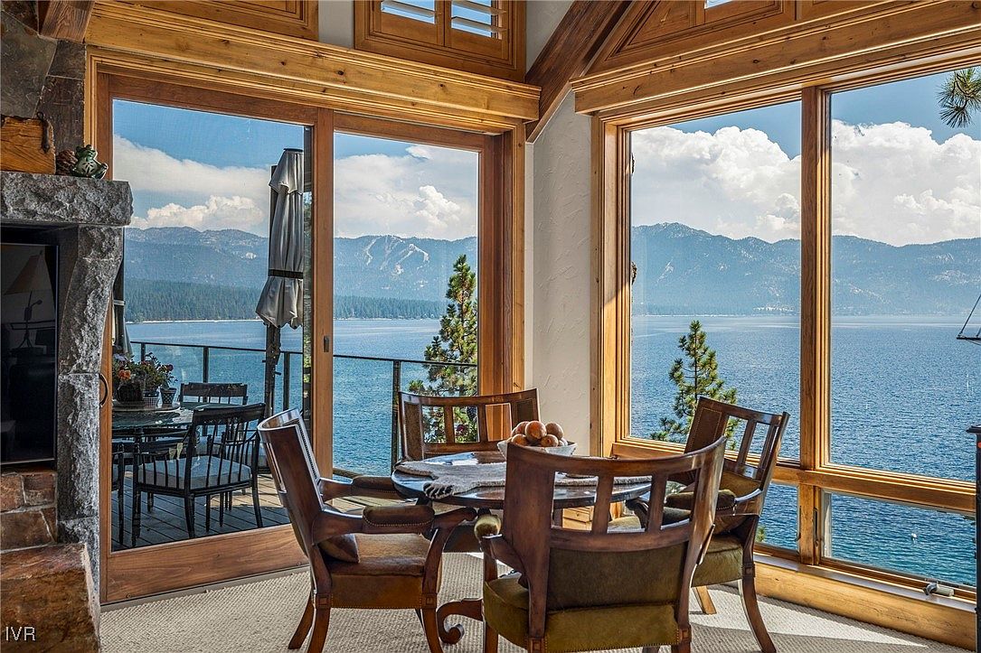 This is an interior shot of a dining room with stunning lake views. The room features a round wooden dining table surrounded by wooden chairs with upholstered seats. Large windows offer panoramic views of the lake and mountains, creating a serene and luxurious dining experience.