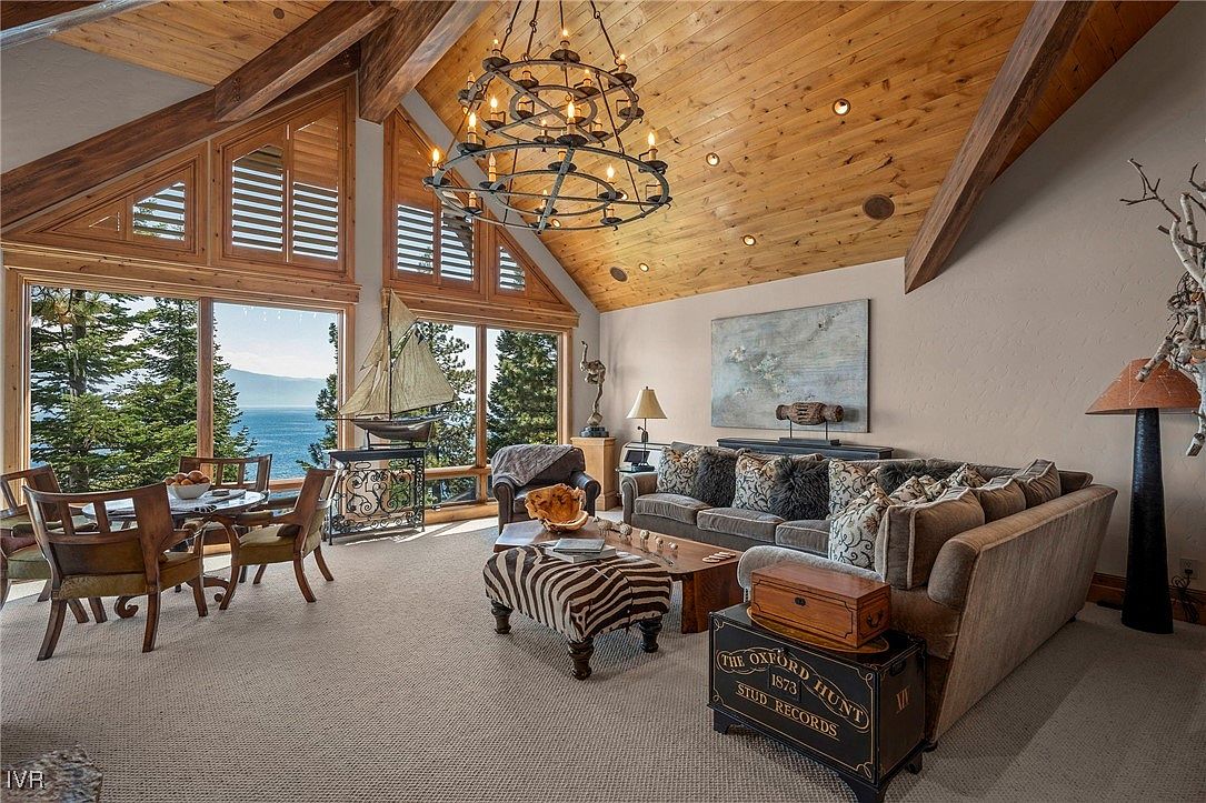 This is an interior shot of a luxurious living room featuring a high vaulted ceiling with exposed wooden beams and a large chandelier. A wall of windows offers a stunning view of the lake and surrounding trees, while comfortable seating and elegant decor create a warm and inviting atmosphere. The room is well-lit and appears spacious, perfect for relaxation and entertaining.