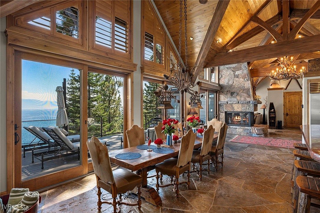 This is an interior shot of a luxurious dining room featuring a large wooden table set for a meal, surrounded by elegant chairs. The room boasts high ceilings with exposed wooden beams and a stone fireplace, creating a warm and inviting atmosphere. Large windows offer a stunning view of the lake and surrounding landscape, enhancing the room's appeal.