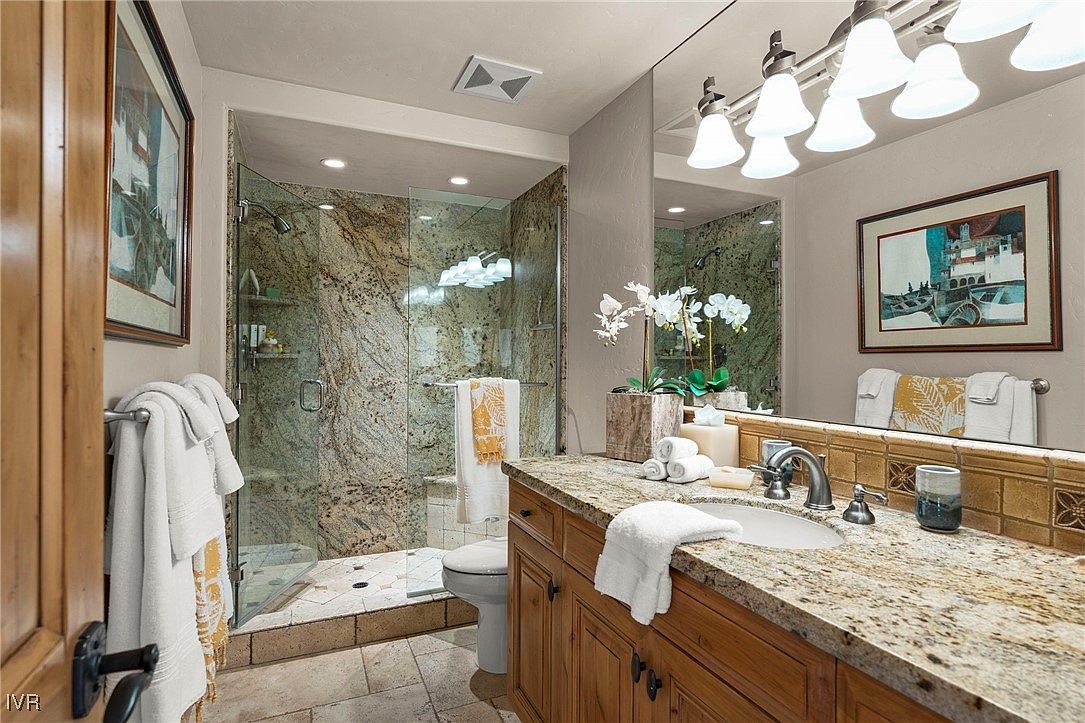 This is a primary bathroom featuring a granite countertop vanity with wooden cabinets, a sink, and a large mirror with decorative lighting. The shower is enclosed with glass and has marble-like walls. The overall style is luxurious and well-maintained, creating a spa-like atmosphere.