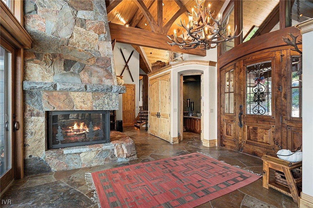 This interior shot showcases a grand entryway featuring a stone fireplace, a large wooden door with decorative glass, and a striking antler chandelier. The flooring is stone tile, complemented by a red patterned rug. The space exudes a rustic, luxurious feel with its exposed wooden beams and natural materials, creating a warm and inviting atmosphere.