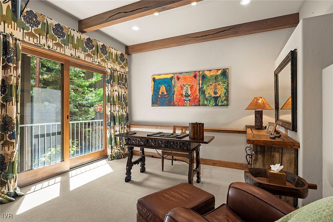 This interior shot showcases a cozy office or study area with a rustic yet refined aesthetic. The room features wooden ceiling beams, a dark wood desk with intricate detailing, and a colorful triptych of bear portraits above it. A sliding glass door leads to a balcony, providing natural light and a view of the surrounding greenery, while patterned curtains add a touch of whimsy.