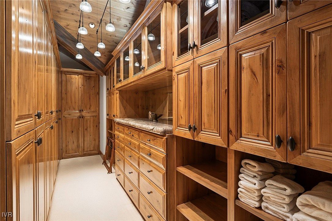 This is a well-organized walk-in closet featuring custom wood cabinetry with glass-fronted upper cabinets and drawers. The closet includes open shelving for towels and accessories, and the warm wood tones create a luxurious feel. The space is illuminated by multiple recessed lights, highlighting the ample storage and organization options.
