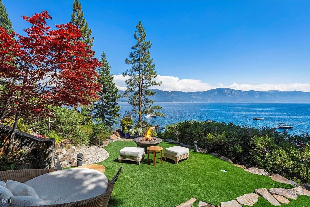This image showcases a beautifully landscaped yard with a stunning view of a lake and mountains. The focal point is a cozy outdoor seating area with a fire pit, surrounded by lush greenery, trees, and decorative stone pathways. The scene evokes a sense of luxury and relaxation, perfect for enjoying the natural surroundings.