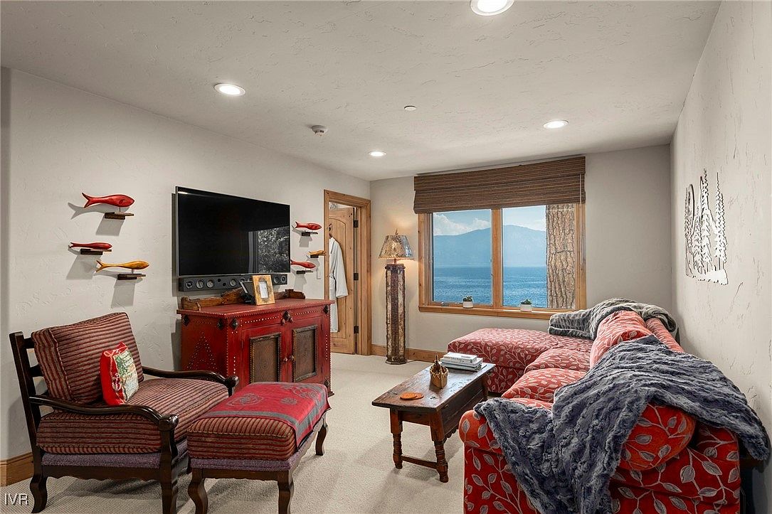 This is an interior shot of a cozy living room featuring a red patterned sofa with a blue throw blanket, a wooden coffee table, and a red cabinet with a flat-screen TV mounted above. The room is decorated with fish-themed wall art and has a large window offering a view of a lake or ocean. The overall impression is warm and inviting.