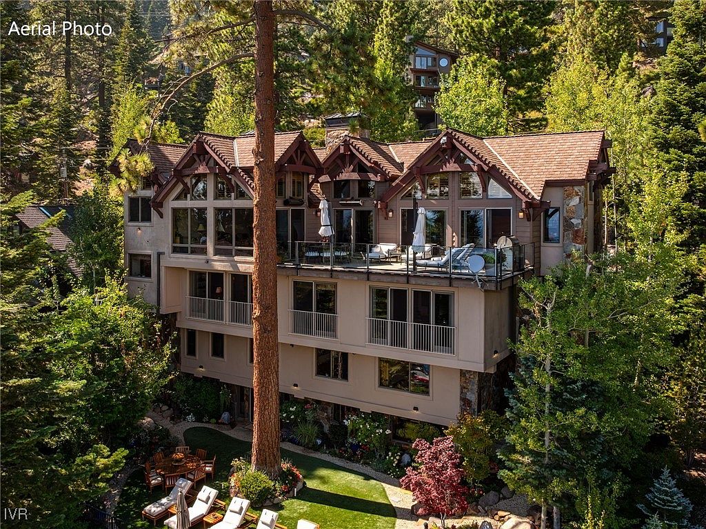 This aerial view showcases a multi-level luxury home nestled among tall trees. The house features a brown roof, multiple balconies, and large windows that offer stunning views. The well-maintained yard includes a green lawn, patio furniture, and colorful landscaping, creating an inviting outdoor space.