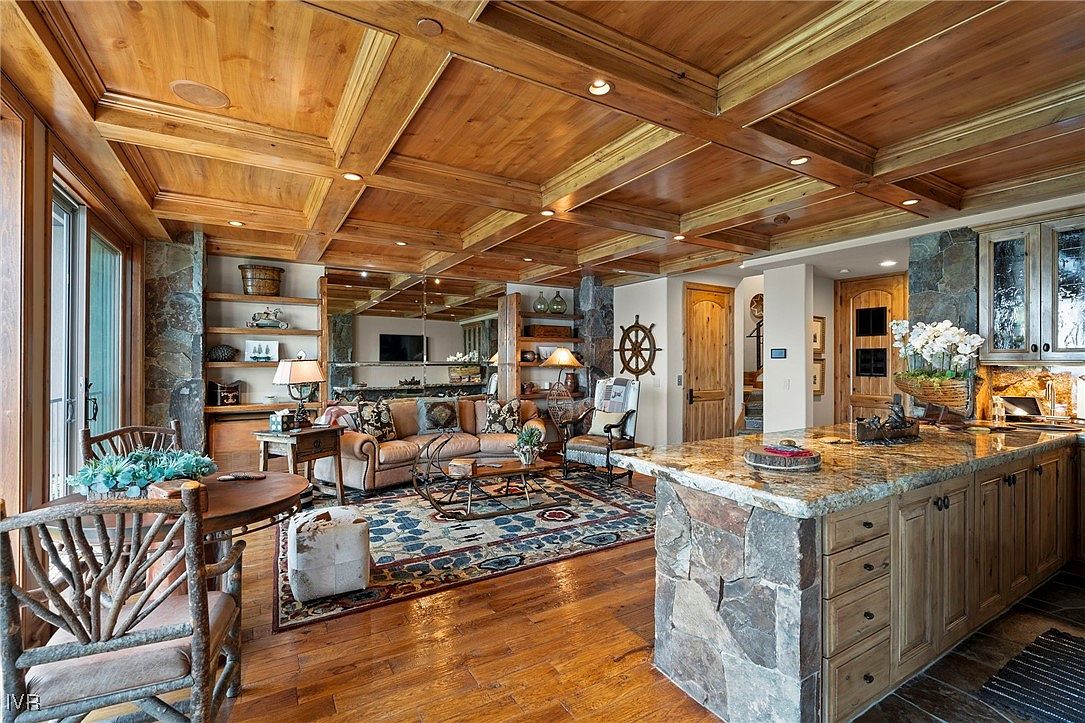 This is an interior shot of a luxurious living room featuring a coffered wood ceiling, hardwood floors, and a stone-accented kitchen island. The room includes a comfortable leather sofa, a decorative rug, and built-in shelving, creating a warm and inviting atmosphere. The space is well-lit and appears to be part of an upscale home.