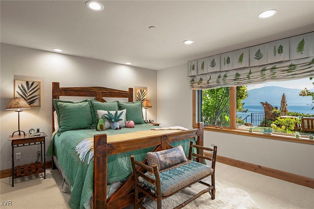 This is a cozy primary bedroom featuring a rustic wooden bed with green bedding and decorative pillows. A window offers a scenic view of the lake and surrounding landscape, enhancing the room's tranquil atmosphere. The room is well-lit with recessed lighting and a bedside lamp, creating a warm and inviting ambiance.