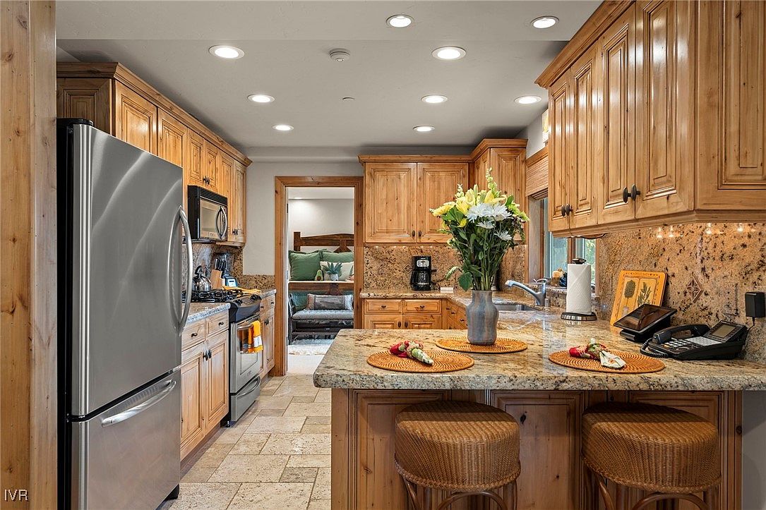 This is a warm and inviting kitchen featuring knotty pine cabinetry and granite countertops. Stainless steel appliances complement the wood tones, while a central island with wicker stools provides a gathering space. The kitchen opens to another room, creating an open and airy feel.