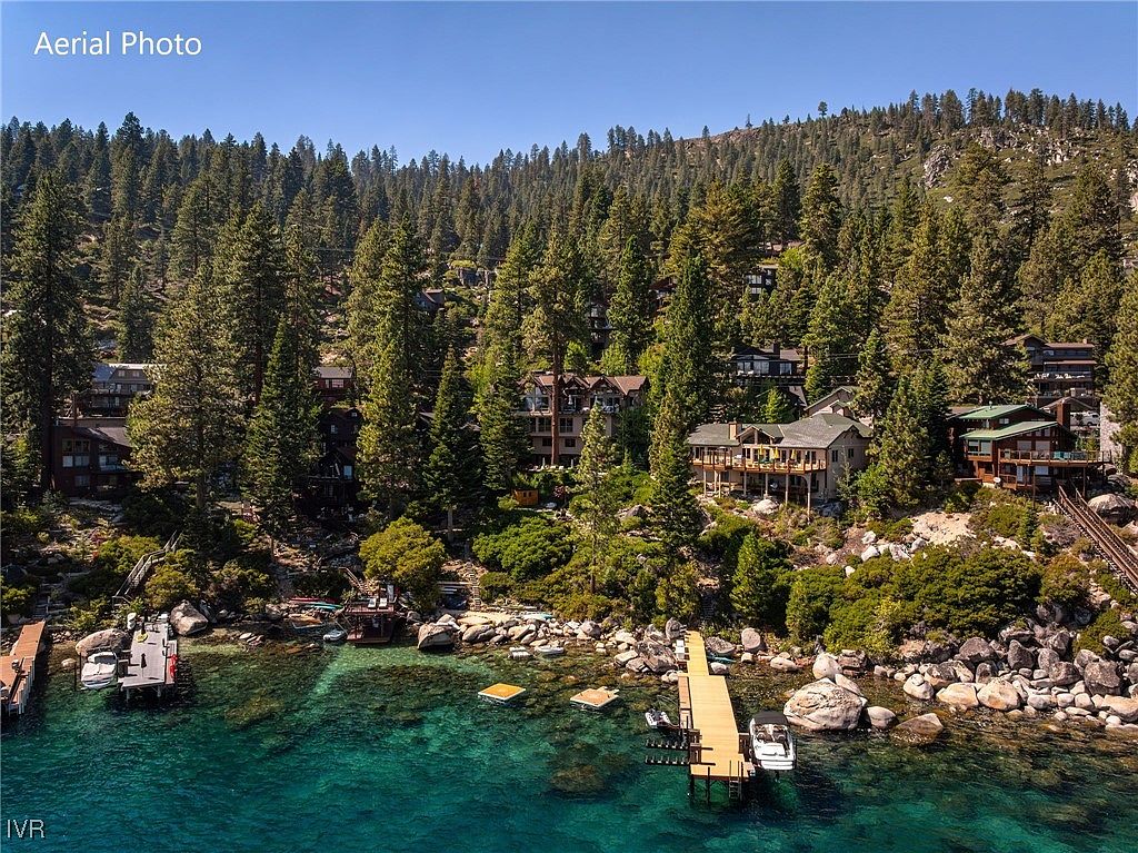 This aerial shot showcases a collection of luxury homes nestled among towering pine trees along a rocky shoreline. The crystal-clear turquoise water reveals the lakebed below, while docks extend into the water, some with boats moored alongside. The scene evokes a sense of secluded, upscale lakeside living.