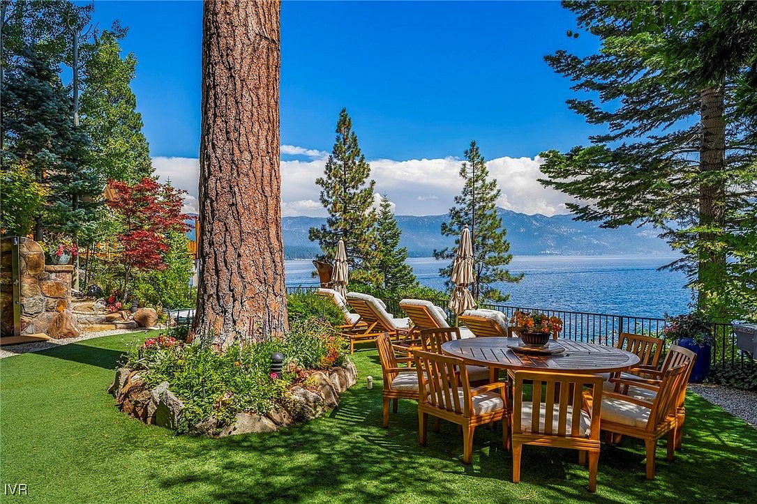 This image showcases a beautifully landscaped yard with a stunning lake view. A round wooden table with chairs sits on a lush green lawn, surrounded by trees and flowering plants. Lounge chairs are also visible, suggesting a relaxing outdoor space perfect for enjoying the scenery.