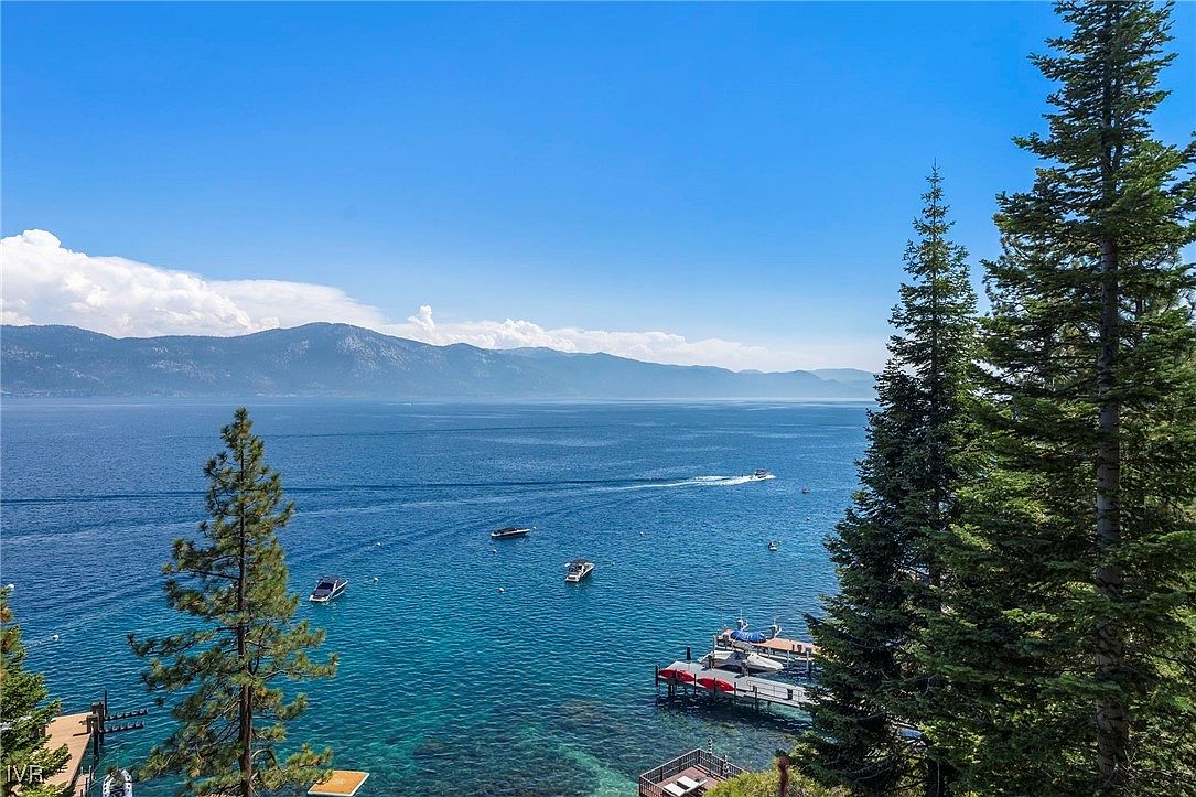 This aerial view showcases a stunning waterfront property with crystal-clear turquoise water and several boats dotting the scene. A private dock extends into the water, offering direct access to the lake. The backdrop features mountains and a clear blue sky, creating a picturesque and serene setting.