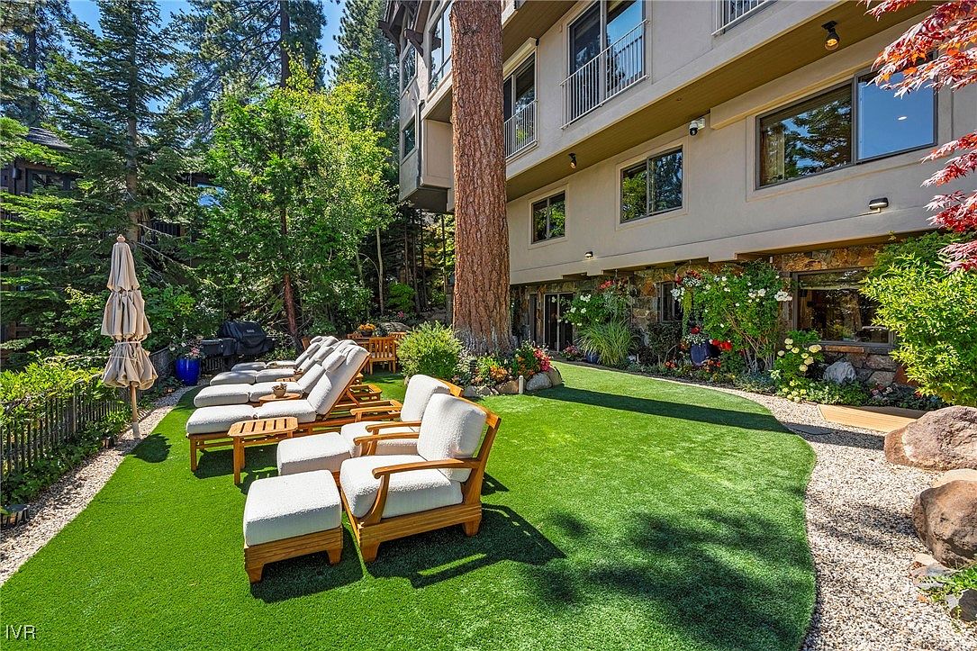 This image showcases a beautifully landscaped backyard featuring a lush green lawn, comfortable outdoor seating, and mature trees. The property's exterior is visible in the background, with windows and balconies adding architectural interest. The scene evokes a sense of relaxation and outdoor living, perfect for entertaining or enjoying quiet moments.