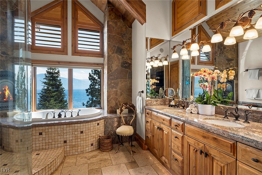This is a luxurious primary bathroom featuring a soaking tub with a view of a lake, a separate shower, and a double vanity with granite countertops and wooden cabinets. The room is decorated with stone accents and features a large mirror and elegant lighting fixtures. The overall impression is one of relaxation and high-end comfort.