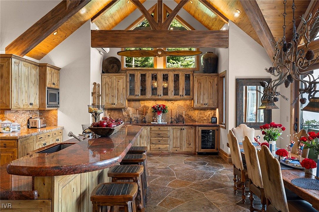 This is a warm and inviting kitchen featuring custom wood cabinetry, granite countertops, and a stone backsplash. The kitchen island provides ample seating, and the open layout connects to the dining area. The rustic design elements, including the wooden beams and stone flooring, create a cozy and luxurious atmosphere.