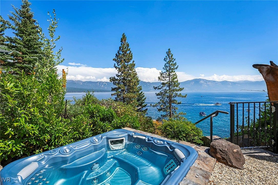 This image showcases an inviting outdoor spa with a stunning view of a lake and mountains. The spa is surrounded by lush greenery and a stone patio, creating a serene and private setting. The clear blue water of the spa contrasts beautifully with the natural landscape, enhancing the property's appeal.