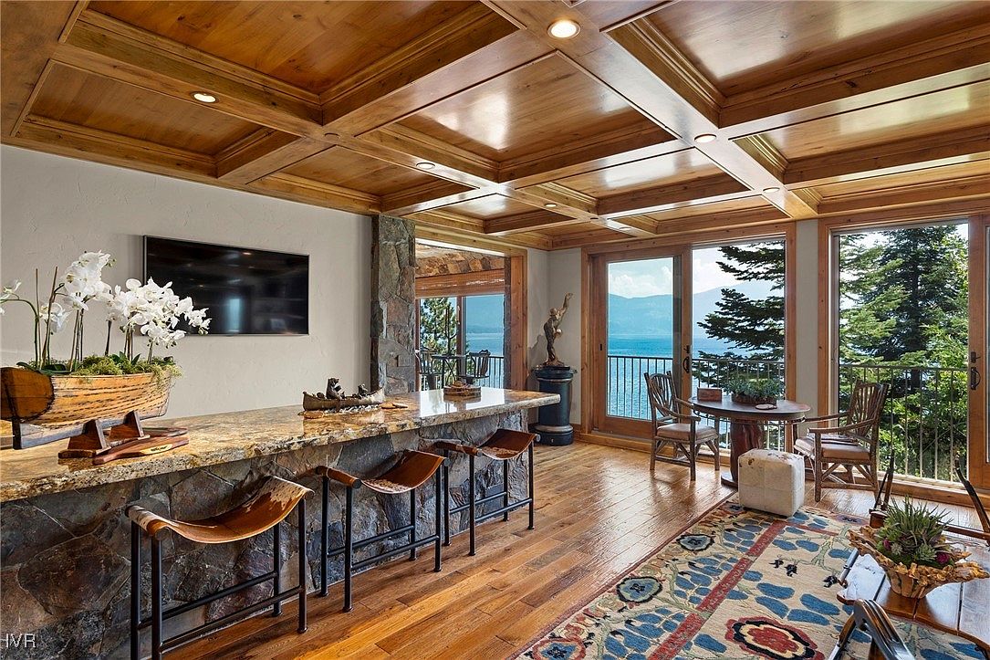 This interior shot showcases a luxurious living room with a stunning lake view. The room features a wooden coffered ceiling, a stone-clad bar with leather stools, and large windows that frame the scenic landscape. The hardwood floors and area rug add warmth and texture to the space, creating an inviting and elegant atmosphere.