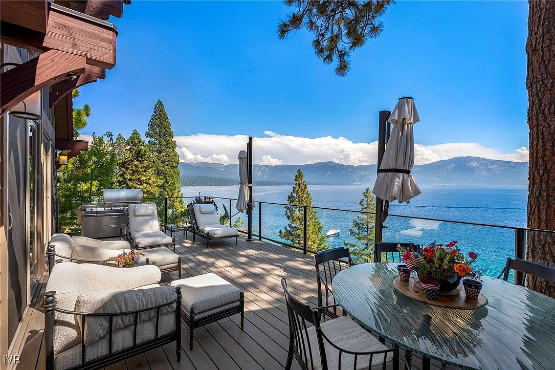 This image showcases a luxurious outdoor deck area with comfortable seating, a grill, and a dining table, all overlooking a stunning lake view. The deck features a glass railing to maximize the view, and the overall impression is one of relaxation and upscale outdoor living. The scene is set on a sunny day with clear blue skies and a calm lake.