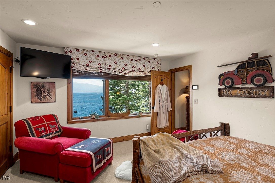 This is a cozy primary bedroom featuring a rustic wooden bed frame with a patterned quilt. A red armchair with a matching ottoman sits near a window offering a scenic view of the water and trees. The room is decorated with a "Ski Country" themed wall hanging and a robe hanging on the door, creating a warm and inviting atmosphere.