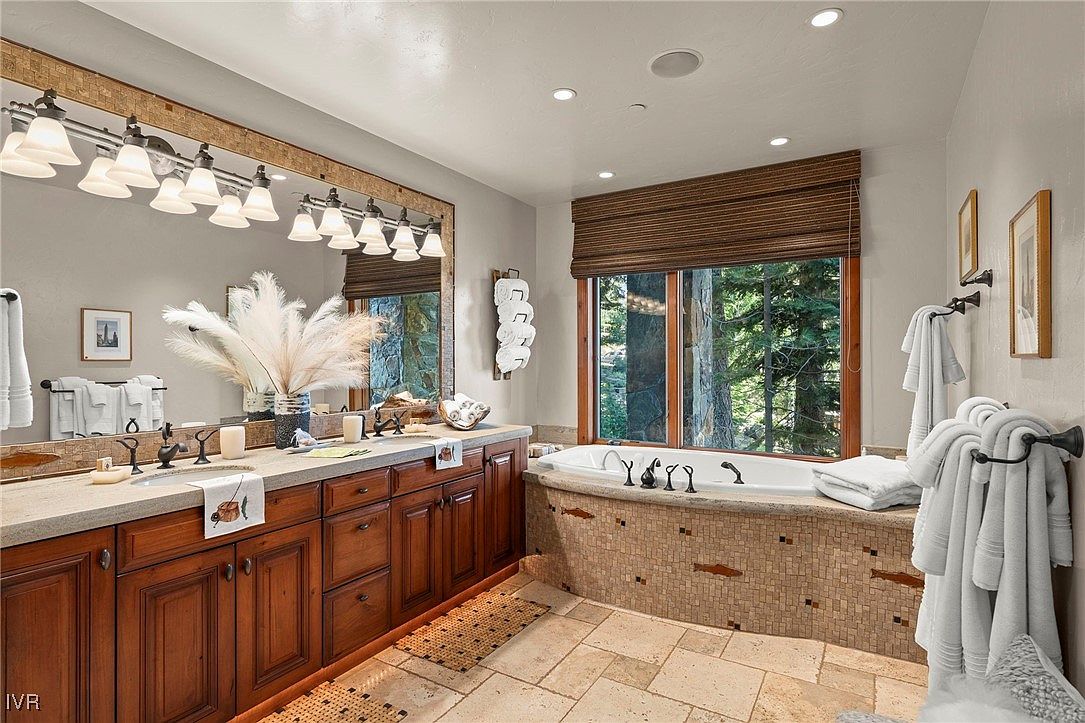 This is a well-appointed primary bathroom featuring a double vanity with dark wood cabinetry and a light-colored countertop. A large mirror with decorative lighting hangs above the vanity. A soaking tub is surrounded by mosaic tile and a large window offering natural light and views of the outdoors. The space is luxurious and inviting.
