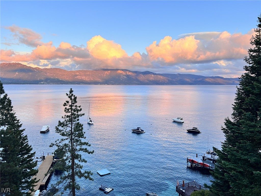 This aerial view showcases a stunning waterfront property with a private dock and several boats on the calm, blue water. The backdrop features mountains under a sky with pink-tinged clouds, creating a serene and luxurious atmosphere. Lush greenery frames the view, adding to the property's appeal and sense of seclusion.
