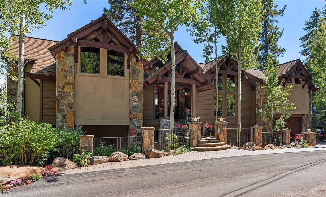 This is a front view of a luxurious mountain home with a rustic yet elegant design. The exterior features a combination of wood siding, stone accents, and exposed timber framing, creating a warm and inviting aesthetic. A well-manicured front yard with a stone and iron fence adds to the property's curb appeal.